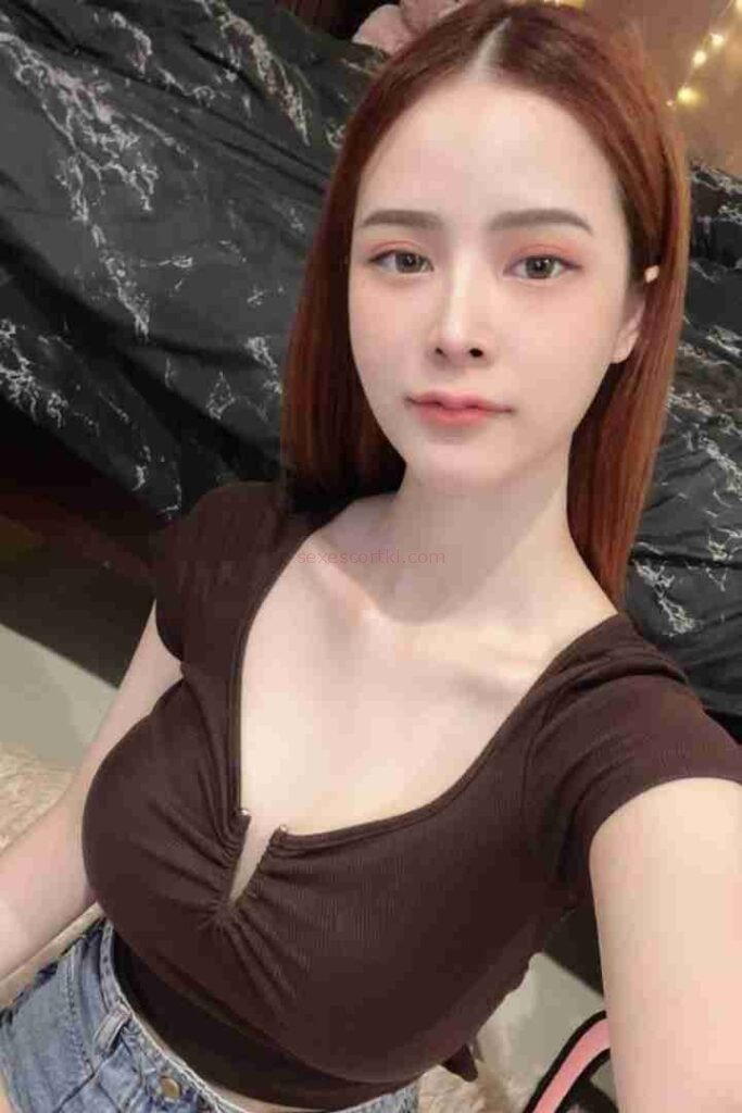 Attractive Asian woman with auburn hair wearing a brown top and jeans. Smiling slightly.