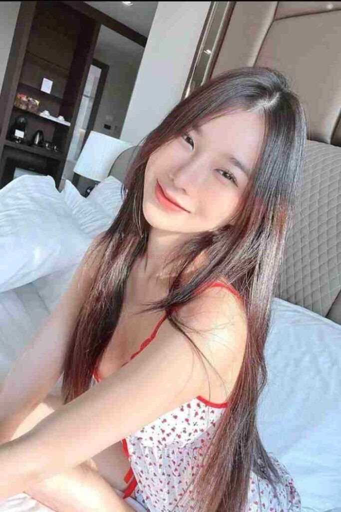 Smiling Asian woman with long hair in patterned top on bed. Ming Yue Chinese Call Girl.