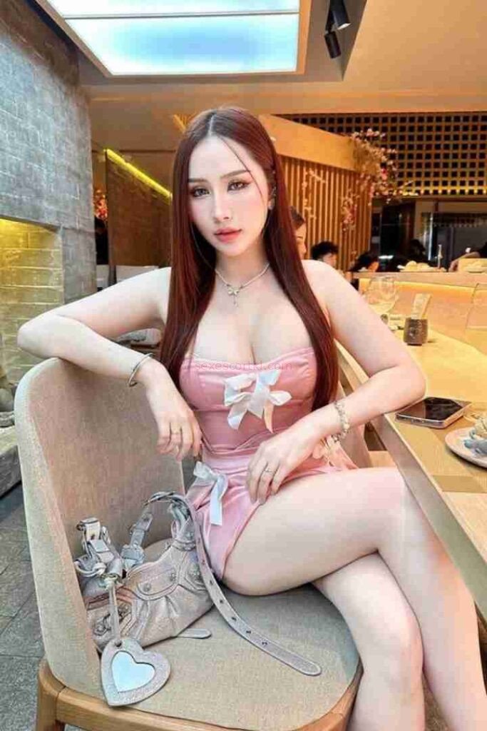 Nin Call Girl in pink dress sitting in chair with purse. Long hair, jewelry, restaurant setting.