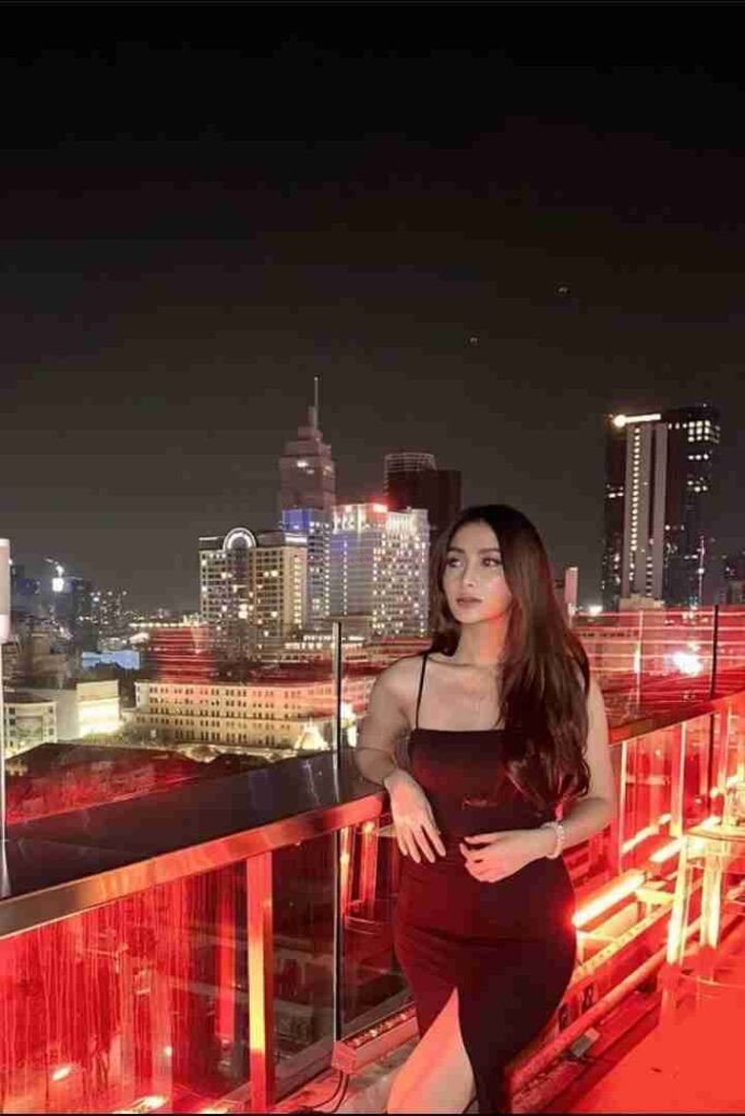 Woman in black dress on rooftop with city lights in background.