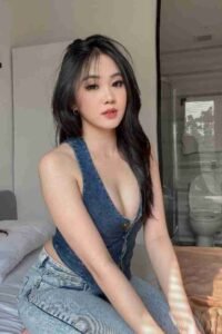Attractive Asian woman in denim vest and jeans sitting on bed. Long dark hair, light makeup, indoor setting.