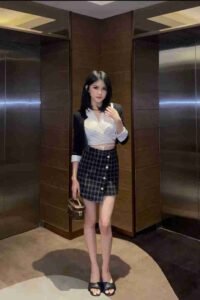 Woman in black blazer, white top, and plaid skirt stands in front of elevators.