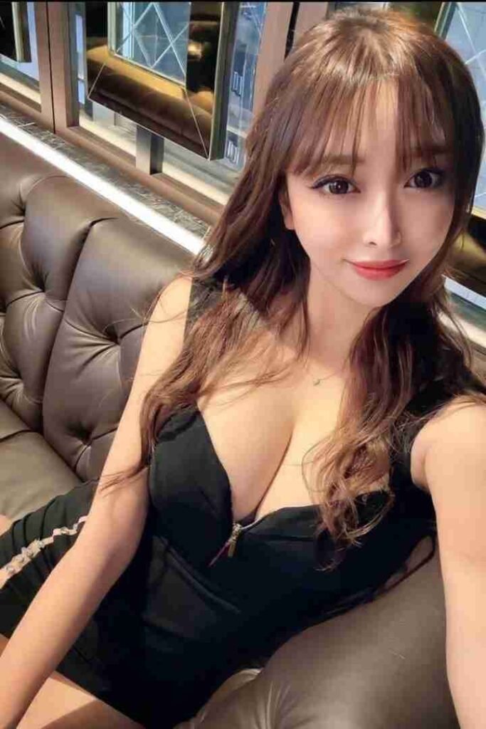 Attractive woman with bangs in black dress sitting on leather couch smiling at camera.