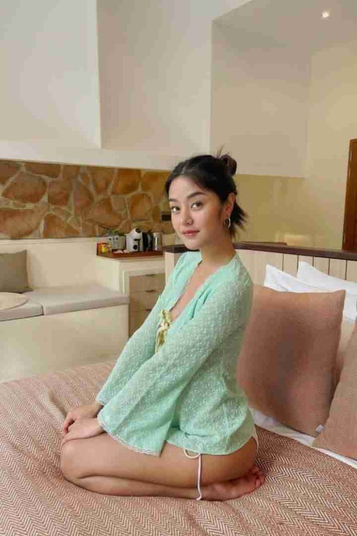 Woman in light green top kneeling on bed, looking at camera. Casual indoor portrait.