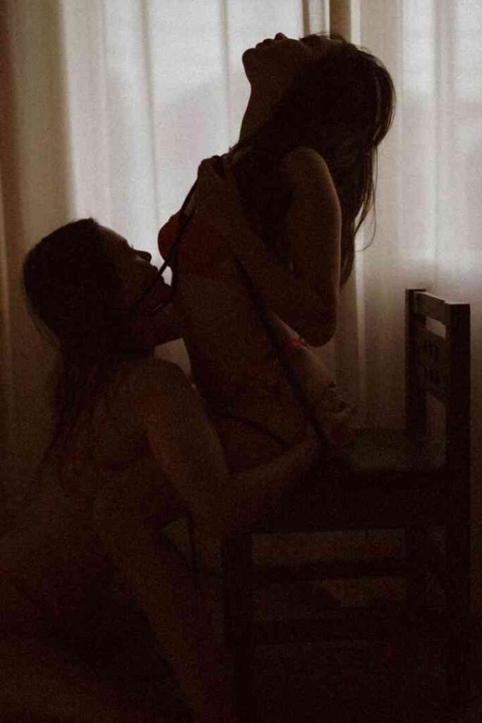 Two women in a dimly lit room, one sitting on a chair and the other kneeling